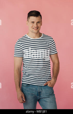 Uomo sorridente in striped t-shirt con la mano in tasca cercando in telecamera isolata su rosa Foto Stock