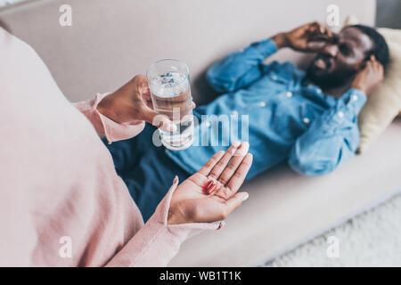 Vista ritagliata della african american donna tenendo un bicchiere di acqua e pillole mentre in piedi vicino all uomo che soffre di mal di testa sul divano Foto Stock