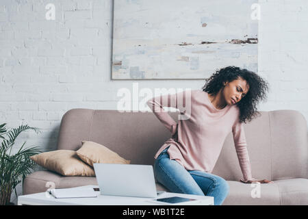 Infelice americano africano donna che soffre di dolori alla schiena mentre è seduto sul divano vicino a tavola con dispositivi digitali Foto Stock