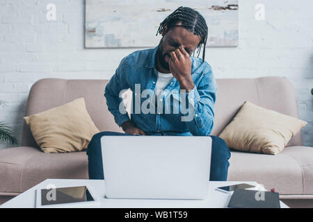 Esaurito americano africano l uomo che soffre di mal di testa mentre è seduto vicino a tavola con dispositivi digitali Foto Stock