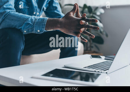 Vista ritagliata della african american uomo seduto con le mani chiuse vicino a tavola con laptop e tablet digitale Foto Stock