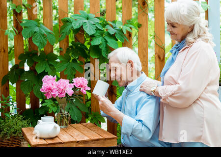 Allegro pensionato donna in piedi vicino a felice uomo senior guardando cup Foto Stock