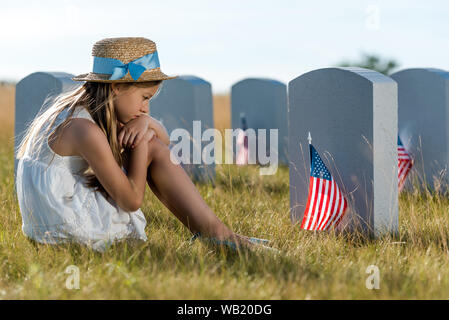 Messa a fuoco selettiva di triste bambino seduto e guardando lapidi con bandierine americane Foto Stock