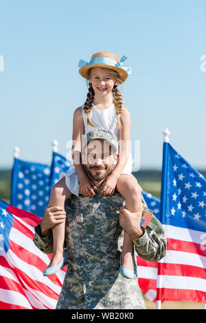 Uomo bello tenendo sulle spalle carino figlia nel cappello di paglia nei pressi di bandierine americane Foto Stock