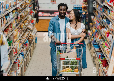 Messa a fuoco selettiva di felice donna asiatica e africana di american uomo a camminare con il carrello nel supermercato Foto Stock