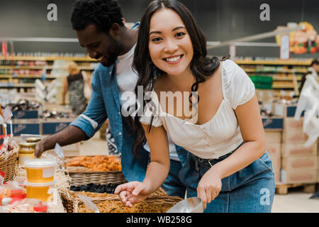 Felice donna asiatica guardando la fotocamera vicino allegro african american uomo guardando barattoli di miele nel supermercato Foto Stock