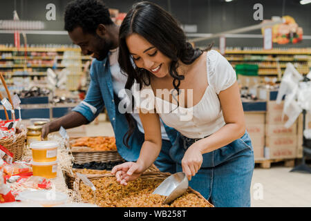 Messa a fuoco selettiva di felice donna asiatica guardando le albicocche secche vicino allegro african american uomo nel supermercato Foto Stock