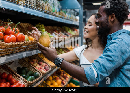 Felice donna asiatica guardando frutti vicino allegro african american uomo in negozio Foto Stock