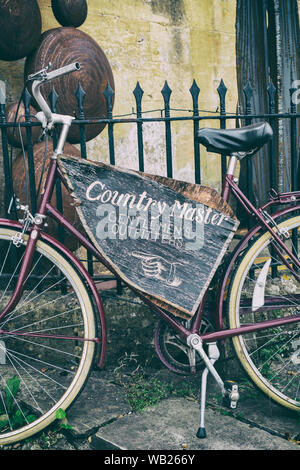 Paese master, Gentlemen's outfitters segno su una bici quaresima contro le ringhiere in Broadway, Cotswolds, Worcestershire, Inghilterra. Vintage filtro applicato Foto Stock