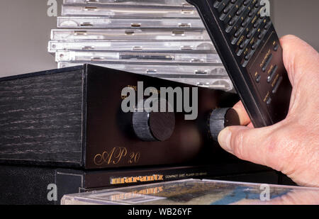 Una mano d'uomo in possesso di un telecomando mentre si ruota una manopola del volume la manopola su un tradizionale analogiche / Amplificatore analogico, con cd impilati. Foto Stock