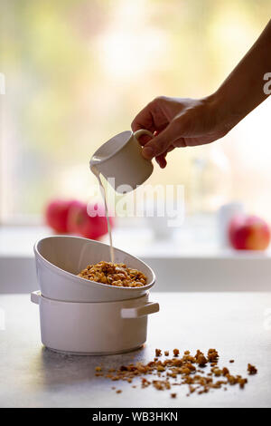 Muesli con noci e uvetta in una ciotola bianco. Una sana colazione la colazione è servita su un tavolo luminoso di fronte alla finestra.Il latte viene colata in una ciotola da una tazza Foto Stock