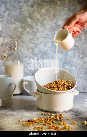 Muesli con noci e uvetta in una ciotola bianco. Il latte viene colata in una ciotola da una tazza. Femmina è mano versando il latte. In verticale Foto Stock