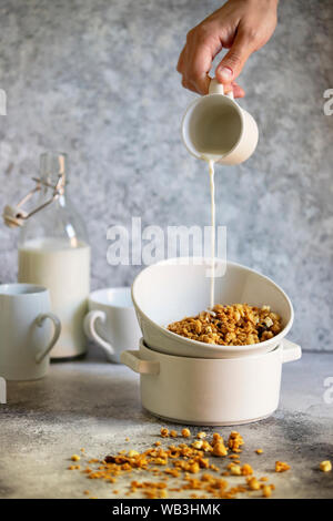 Muesli con noci e uvetta in una ciotola bianco. Il latte viene colata in una ciotola da una tazza. Femmina è mano versando il latte. In verticale Foto Stock