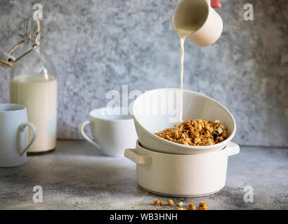 Muesli con noci e uvetta in una ciotola bianco. Il latte viene colata in una ciotola da una tazza. Femmina è mano versando il latte. Luce toni neutri Foto Stock