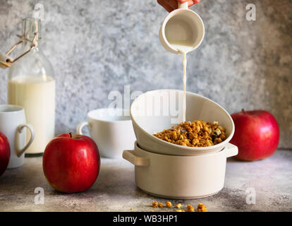 Muesli con noci e uvetta in una ciotola bianco. Il latte viene colata in una ciotola da una tazza. Femmina è mano versando il latte. Luce toni neutri Foto Stock