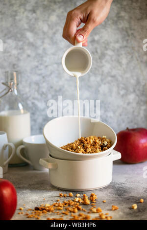 Muesli con noci e uvetta in una ciotola bianco. Il latte viene colata in una ciotola da una tazza. Femmina è mano versando il latte. In verticale Foto Stock