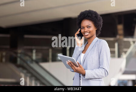 Giovane donna parlando al cellulare, utilizzando digitale compressa vicino ufficio moderno edificio Foto Stock