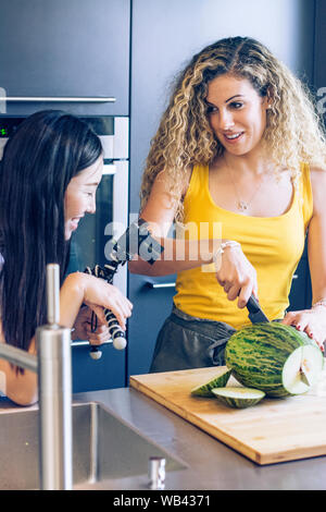 Giovane ragazza bionda il taglio di un melone con un coltello in cucina, mentre un altro record ragazza lei con una telecamera Foto Stock