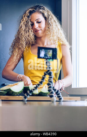 Giovane ragazza bionda il taglio di un melone con un coltello da cucina, durante la registrazione con una telecamera Foto Stock