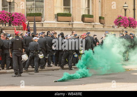 Portland Place, Londra, Regno Unito. 24 agosto 2019. Gli agenti della polizia metropolitana si riuniscono in una strada di Londra in mezzo a un fuoco di fumo verde durante una dimostrazione pubblica. Gli ufficiali in uniforme nera e caschi si trovano vicino a bandiere ed edifici ricoperti di fiori, suggerendo il controllo della folla o il coordinamento tattico. Protesta libera di Tommy Robinson fuori dalla BBC. Allo stesso tempo si sta svolgendo una contromanifestazione, organizzata da Stand Up to razzism and Unite Against fascism. Penelope Barritt/Alamy Live News Foto Stock