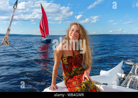 Giovane donna sorridente con i suoi capelli disheveled dal vento è seduta sul bordo di un catamarano a vela sullo sfondo di paesaggi acquatici con uno yacht Foto Stock