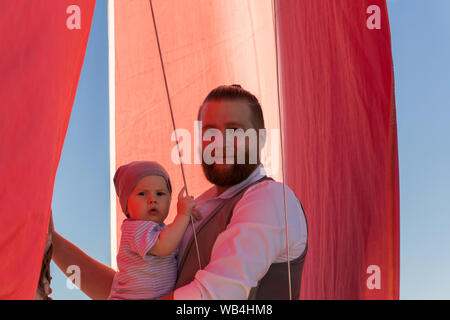 Uomo Barbuto tenendo un bambino nelle sue braccia contro lo sfondo del cielo blu e un montante di nave con vele di rosso Foto Stock