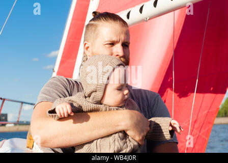 Uomo Barbuto tenendo un bambino nelle sue braccia contro lo sfondo del cielo blu e un montante di nave con vele di rosso Foto Stock