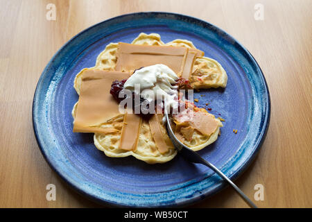 Formaggio norvegese marrone su un waffle con marmellata di fragole e panna acida parzialmente mangiato con una forchetta Foto Stock