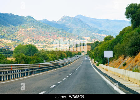 Strada vuota senza auto sull'Isola di Sardegna in Italia in estate. Panorama con autostrada e il verde della natura e il blu del cielo. Le montagne sullo sfondo. Foto Stock