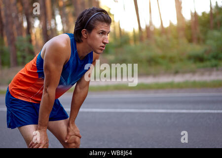 Runner uomo riposo dopo la formazione in esecuzione per il triathlon. Maschio modello fitness triatleta e rilassante. Jogging uomo prendendo una pausa durante il training outdoo Foto Stock