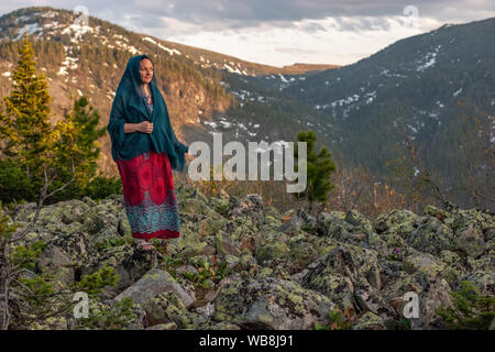 Bella ragazza carina con un vestito e un foulard sorge su grandi pietre in montagna. Contro lo sfondo delle montagne con la neve ricoperta con fores Foto Stock