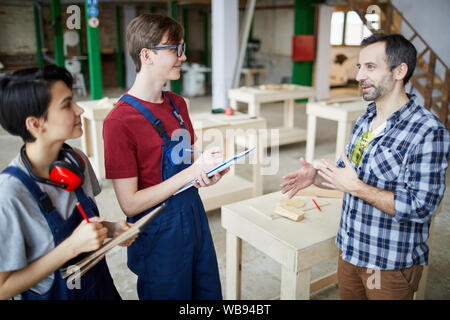 Vista laterale ritratto della coppia carpenter insegnamento gruppo di partecipanti tenendo cartelline in falegnameria officina, spazio di copia Foto Stock
