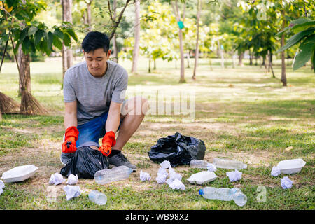 Giovane uomo asiatico indossando guanti di colore arancione e il cestino di raccolta nel sacchetto di immondizia nel parco. Salvare la terra e le preoccupazioni ambientali concetto Foto Stock