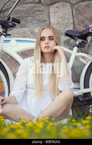 Triste bella ragazza bionda in abito bianco con la bici nei pressi di appoggio della parete, seduta in erba con fiori Foto Stock