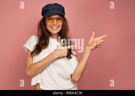 Una bruna in una T-shirt bianco, blu cappello e occhiali da sole sorride ampiamente i punti e le dita al lato su uno sfondo rosa Foto Stock