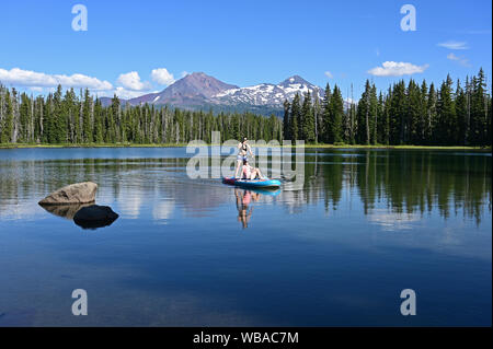 Due giovani donne su standup paddle board sul Lago di Scott, Oregon con il centro e il nord di Sorelle vulcani in background. Foto Stock