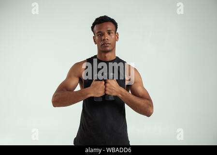 Ottimo allenamento. Muscoloso giovane africano nero holding manubri e guardando la telecamera mentre in piedi contro uno sfondo grigio. Studio shot. Sport concept Foto Stock