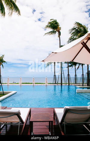 Ago 6, 2014 Hua Hin, Thailandia - mare tropicale e in stile resort piscina infinity edge e con lettino in spiaggia sotto ombrellone bianco e alberi di noce di cocco in estate Foto Stock