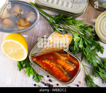 Conserve di pesce e frutti di mare, aprire le lattine con assortimento di conserve di pesce e vongole servita su un tavolo di legno con prezzemolo e pepe Foto Stock