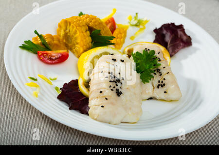 Cod di 250 grammi preparata in padella con acqua e olio d'oliva per 10 minuti. Aggiungere sale e pepe. Serviti con mais bollito, verdi e limone in corrispondenza della piastra Foto Stock