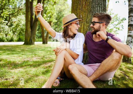 Coppia giovane tenendo selfie seduti contro albero nel parco e guardando ogni altro Foto Stock