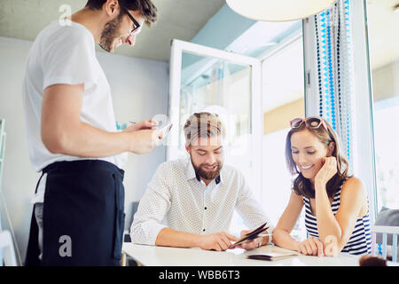 Due giovani ordinare scegliendo dal menu e parlando con cameriere mentre sedendo nel moderno ristorante Foto Stock