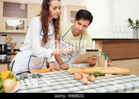 Giovani caucasici in coppia cinghiette ricetta di lettura nel tablet pc prima di cucinare la cena insieme Foto Stock