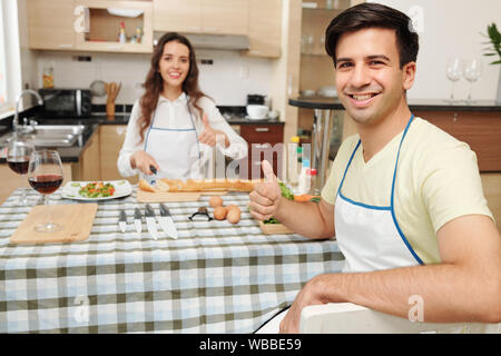Felice giovane coppia caucasica cucinare la cena in cucina e mostrando pollice in su Foto Stock