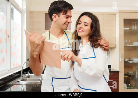 Bel giovane abbracciando la moglie e mostrando la sua nuova ricetta interessante nel libro di cucina Foto Stock