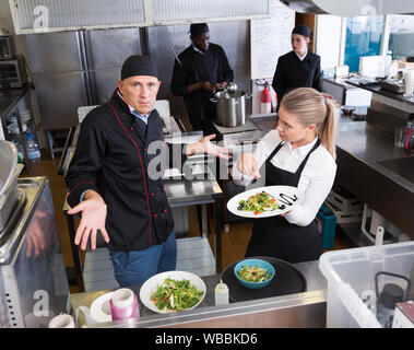 Disorientati chef rendendo impotente gesto mentre parla con sorpresa la cameriera che puntano a piatto cucinato Foto Stock