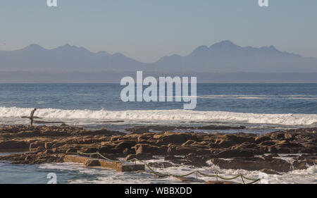 Mossel Bay, Sud Africa - Un misterioso holiday maker esplora le rocce a La Punt Beach con un impianto di trivellazione petrolifera nella baia in background Foto Stock