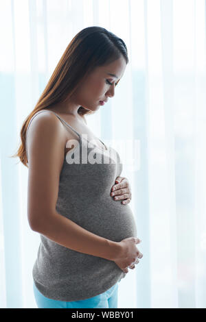 Calma giovane vietnamita donna incinta in piedi contro la finestra e toccando il suo ventre Foto Stock