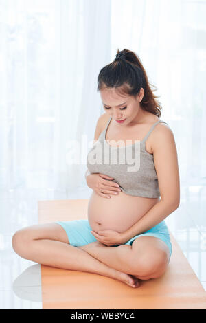 Sporty giovani asiatici incinta Donna seduta sul materassino yoga e toccando il suo grande ventre con amore e cura Foto Stock