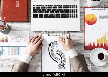 Vista dall'alto ufficio moderno spazio di lavoro con mano umana schizzo su un foglio di carta. Piatto bianco laici scrivania in legno con la tastiera portatile, business infographics e tazza di Foto Stock